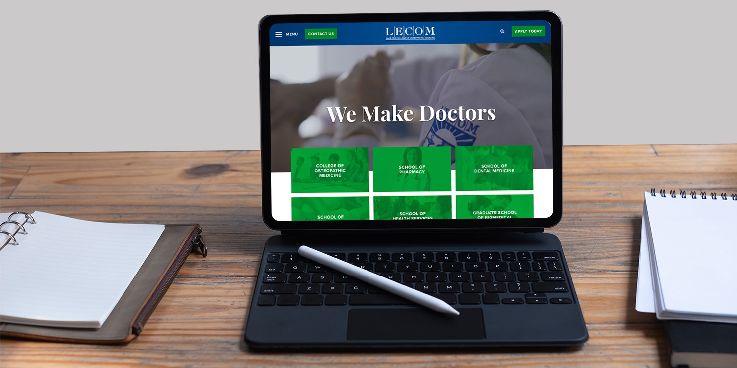 LECOM.edu website on tablet laptop on wooden desk with notebooks and pens surrounding.